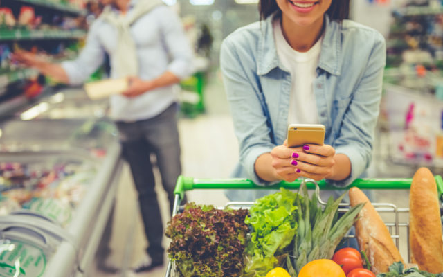 A cropped image of a woman leaning on a shopping cart, in a grocery stores, while she is using a mobile phone and smiling. Inn the background is a man shopping for food. Photo from Alpha Media USA Portland OR