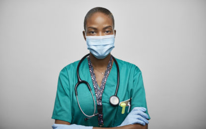 Photo of a female healthcare worker wearing a blue medical face mask, blue latex gloves, teal colored scrubs and a stethoscope. Photo from Alpha Media USA Portland OR